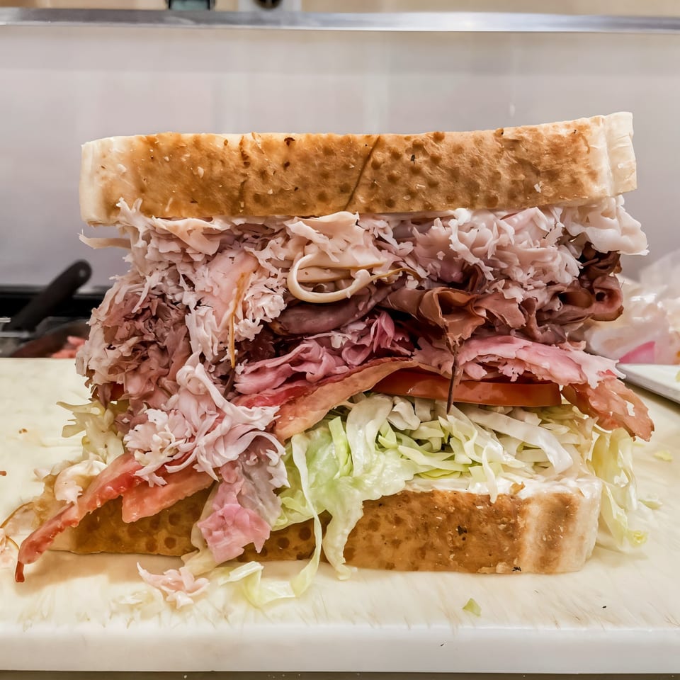 A massive, overstuffed sandwich with various deli meats, lettuce, and tomatoes on thick-cut bread.