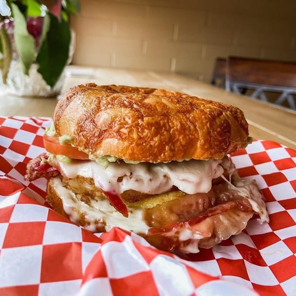 A bagel sandwich with melted cheese, bacon, and tomato on red and white checkered paper.