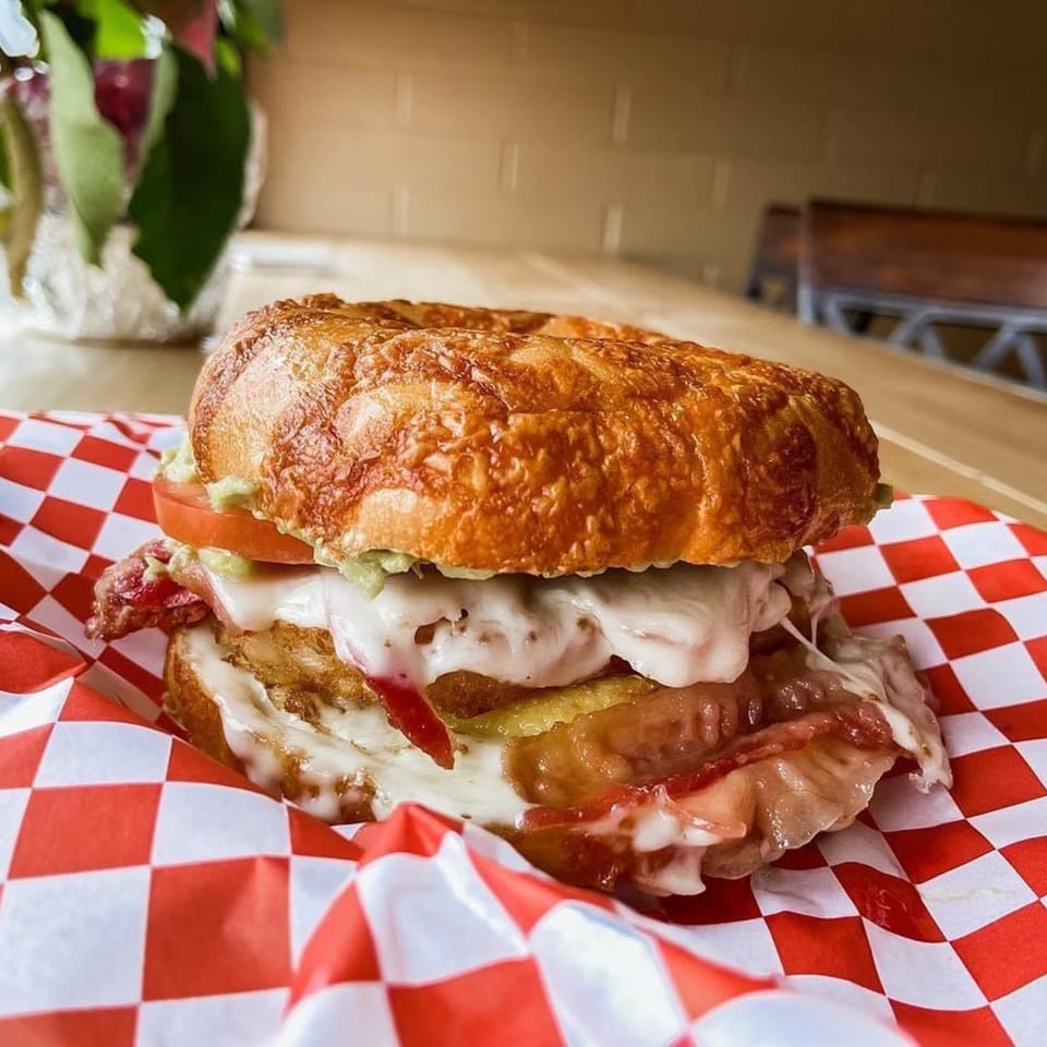 A bagel sandwich with melted cheese, bacon, and tomato on red and white checkered paper.
